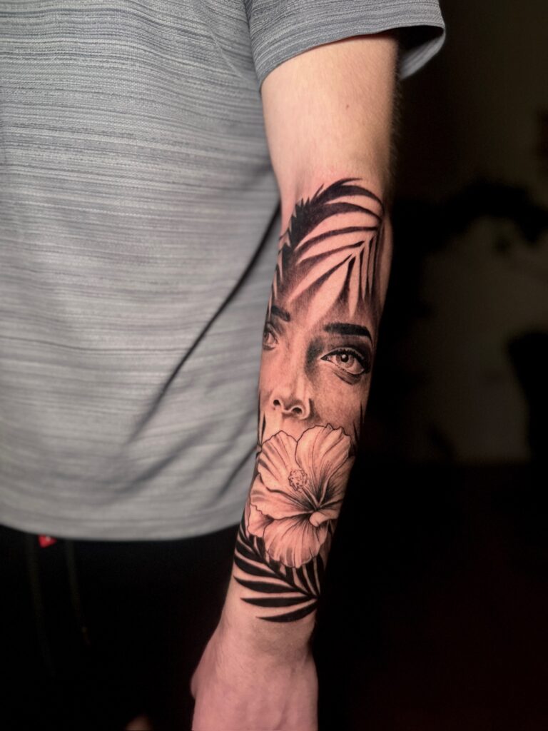 Black and grey forearm tattoo combining a portrait, flower and foliage